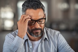 Frustrated middle aged man sitting on couch at home. Close up face of stressed indian businessman wearing eyeglasses with eyes closed. Overworked middle eastern business man with terrible migraine.