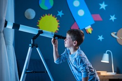 Curious boy using telescope to explore moon surface at night in his bedroom. Young child using telescope to see remote galaxy from room with decorated wall with rocket, planets, stars and spaceship.