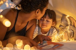 African american mother kissing her daughter on forehead while lying on bed in illuminated tent. Mother putting daughter to sleep in cozy hut after reading a fairy tale in tent. Mom kissing cute girl.