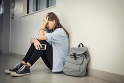Upset and depressed girl holding smartphone sitting on college campus floor holding head. University sad student suffering from depression sitting on floor at high school. Lonely bullied teen.