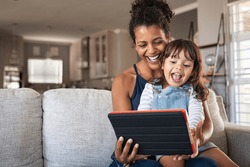 Young black mother and smiling daughter playing on digital tablet at home with copy space. Young african american woman with cheerful and excited little girl using digital tablet on couch at home.
