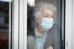 Lonely old woman wearing surgical mask and looking through the window during lockdown. Senior sad woman with face protective mask stay at home. Depressed lady at home during the covid-19 pandemic.