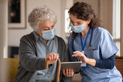 Doctor and senior woman going through medical record on digital tablet during home visit wearing face mask. Old woman with nurse with surgical mask and using digital tablet during coronavirus pandemic
