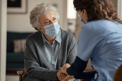 Elderly woman talking with a doctor while holding hands at home and wearing face protective mask. Worried senior woman talking to her general practitioner visiting her at home during virus epidemic. 