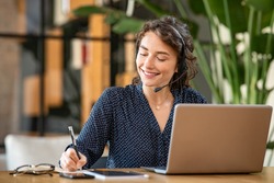 Successful young business woman working on laptop with headphones in a call center as a consultant. Happy businesswoman with headset translating and writing notes by listening to audio course.