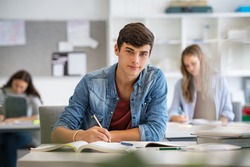 Happy student taking notes while studying in high school. Satisfied young man looking at camera while sitting at desk in classroom. Portrait of college guy writing while completing assignment.