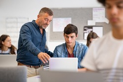 Professor assisting college student with laptop in classroom during computer lesson. Teacher talking and explaining to guy. Mature man lecturer helping high school teen with laptop during lecture.