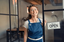 Portrait of positive business woman standing at cafeteria door entrance. Cheerful young waitress in blue apron near glass door with open signboard and looking at camera. Excited small business owner.