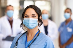 Close up face of confident female nurse in front of his medical staff looking at camera while wearing protective face mask due to covid-19 virus. Smiling surgeon standing  with team in background.