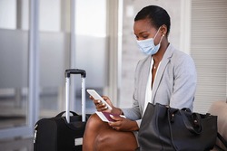 Mature black businesswoman using mobile phone at the airport in the waiting room while wearing face mask due to covid-19. African business woman holding passport and boarding pass typing on smartphone