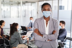 Successful black entrepreneur in conference room leaning over table wearing surgical mask with business people working in background. Portrait of african american leader woman looking at camera.