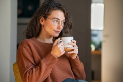 Pensive woman drinking hot coffee at home. Thoughtful young woman drinking a cup of tea while thinking. Pretty contemplative girl with sweater relaxing at home while drinking purifying herbal tea.