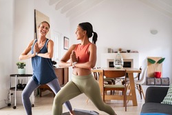 Mature woman practicing yoga at home with mixed race friend. Beautiful sporty indian woman with female friend in yoga position of the warrior at home. Two middle aged lady exercising at home, lockdown