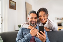 Mid adult man relaxing on sofa and showing new app to african american wife on cellphone. Middle eastern man and woman sitting on couch at home and using mobile phone to do a video call with family.