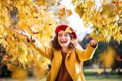 Beautiful young woman throwing autumn yellow leaves. Portrait of joyful woman playing with leaves in park during fall. Happy girl wearing coat and red beret playing with yellow leaves outdoor.