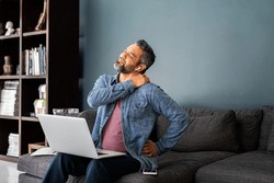 Mature indian man suffering from shoulder and back pain while sitting on couch and working from home on laptop. Stressed middle eastern businessman suffering from neck pain and stretching.