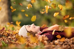 Cheerful young woman lying over leaves with closed eyes and smiling. Woman lying over autumn ground relaxing at park. Casual joyful girl having fun throwing leaves in autumn park with copy space.