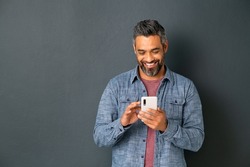 Mid adult multiethnic man texting phone message on smart phone isolated on grey background. Smiling middle eastern man using smartphone on gray wall. Happy mixed race guy using app on mobile phone.
