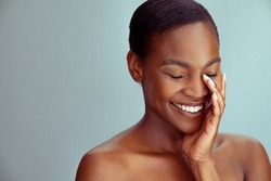 Smiling mature african woman with bare shoulder and eyes closed isolated against light blue wall. Beautiful mid african american lady with fresh glowing skin and bare shoulder looking down.