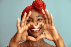 Close up of hands of beautiful african woman making heart shape isolated against blue wall. Portrait of smiling middle aged black woman making heart gesture with hands around her big white smile.