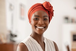 Portrait of smiling middle aged African american woman with headscarf at home. Mid adult black woman with turban looking at camera. Happy mid mature lady wearing traditional african scarf on head.