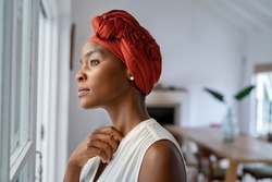 Pensive woman wearing headscarf looking outside window in contemplation. Mature black woman wearing a traditional turban and thinking near window at home. Worried african mature woman with cancer.