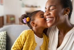 Close up of beautiful daughter kissing mother on cheek at home. African little girl giving kiss to happy mother. Lovely black female child kissing cheerful and proud woman on cheek for mother's day.