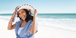 Portrait of stylish latin hispanic woman with white straw hat standing at beach. Young smiling woman on vacation enjoy sea breeze wearing straw hat and looking at camera. Attractive beautiful girl.