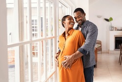Lovely mid black man hugging his pregnant wife from behind standing near window at home. Happy middle aged black husband embracing pregnant woman while waiting for baby. African american mature couple