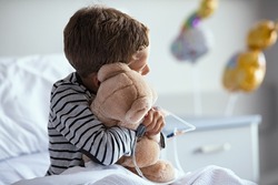 Rear view of scared little boy with intravenous drip in hand hugging teddy bear sitting on hospital bed. Sick lonely child looking through the window in clinic pediatric ward before the surgery.