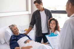 Happy senior woman lying on hospital bed with lovely son and daughter visiting and talking to doctor. Professional physician gives the results of the medical report to patient's family members.