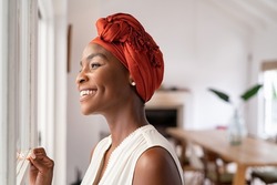 Smiling mid adult african american woman with turban looking outside the window. Cheerful black mature woman wearing traditional headscarf while contemplating. Happy middle aged lady thinking.