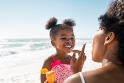 Young mother applying protective sunscreen on daughter nose at beach with copy space. Black woman hand putting sun lotion on female child face. African american cute little girl with sunblock cream.