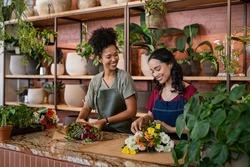 Two happy and smiling young woman florist working together in flower shop. Florist talking and making beautiful bouquet of flower together with her black colleague. Beautiful florists arranging bunch.