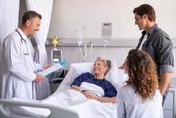 Mature happy doctor giving good news to family and old patient. Smiling senior woman lying on hospital bed with son and daughter visiting and talking to physician. Doctor visiting hospitalized patient