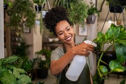 Beautiful black florist taking care of plants while spraying it with water. African american owner working and spraying water plants in store. Happy and smiling florist watering plants in shop.
