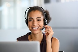Call center agent with headset working on support hotline in modern office. Young african american agent in conversation with customer over headset looking at camera. Portrait of black girl working.