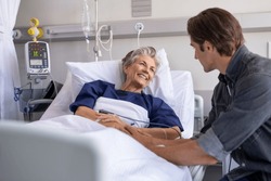 Son visiting sick old mother in hospital. Smiling senior patient in conversation with man. Grandson visiting and cheering his happy granny lying in bed at modern hospital ward.