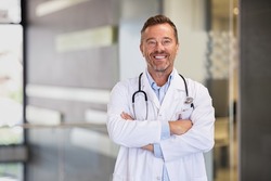 Portrait of happy mature doctor with folded arms standing at hospital hallway. Confident male doctor in a labcoat and stethoscope looking at camera with satisfaction. Smiling general practitioner.