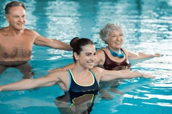 Smiling fitness class doing aqua aerobics in swimming pool. Smiling young woman with senior couple stretching arms in swimming pool. Fit mature man and old woman exercising in swimming pool.