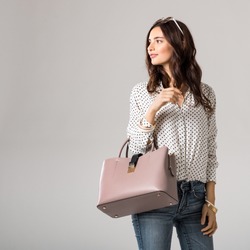 Young glamour woman wearing polka dot shirt and jeans posing with pink handbag. Beautiful stylish girl holding bag and looking away with copy space. Fashion woman holding peach bag with sunglasses.