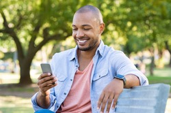 Young african man sitting at park and using smart phone. Happy smiling guy reading a message on cellphone while sitting on a bench in a park. Black man surfing net with smartphone.