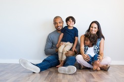 Happy multiethnic family sitting on floor with children. Smiling couple sitting with two sons and looking at camera. Mother and black father with their children leaning on wall with copy space.