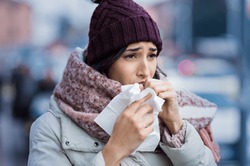 Young woman coughing during winter on street. Girl with cold wearing knitted cap and scarf feeling unwell. Woman feeling sick during for winter and city pollution. Girl with sore throat.