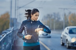 Young fitness woman runner checking time from smart watch. Young woman checking heart rate while jogging on street at dusk. Young woman looking on smartwatch her heartbeat while running in city. 
