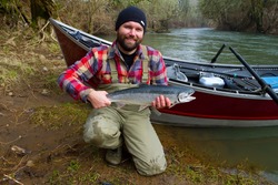 A steelhead fisherman holds his trophy fish by his boat and the river in Oregon.
