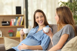 Happy pregnant woman talking with a friend sitting on a couch in the living room at home