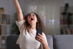 Excited woman celebrating success holding mobile phone sitting on a couch in the night at home