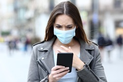 Front view portrait of a scared girl wearing protective mask avoiding contagion reading news on her smart phone on a city street