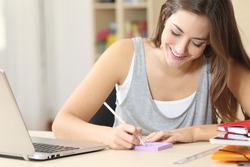 Happy student woman with laptop writing on post note sitting on a desk at home